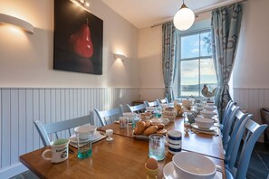 Un coin repas avec une table en bois dressée pour le repas, des chaises, une grande fenêtre avec des rideaux et un luminaire fixé au mur.