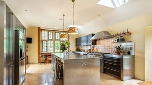 A spacious kitchen with a central island, stainless steel appliances, and a large window providing natural light. - Rissington Hill House (Great Rissington)