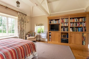 A bedroom with a large bed, a TV, a bookshelf, and a window with curtains. - Rissington Hill House (Great Rissington)