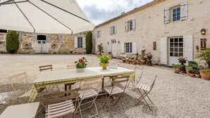 Une cour avec une table et des chaises placées sous un grand parasol, entourée d’un bâtiment doté de nombreuses fenêtres et portes.