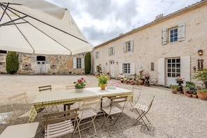 Une cour avec une table et des chaises placées sous un grand parasol, entourée d’un bâtiment doté de nombreuses fenêtres et portes.
