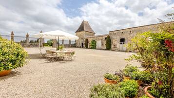 A courtyard with a table and chairs under a canopy, surrounded by stone buildings and potted plants.