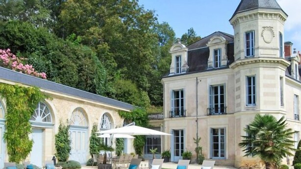 Un espace piscine aménagé avec des chaises longues et une table, entouré d’un grand bâtiment doté d’une tour.