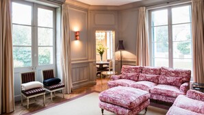 Une pièce dotée de grandes fenêtres, d’un canapé, de deux fauteuils, d’une table avec des chaises et d’une lampe.