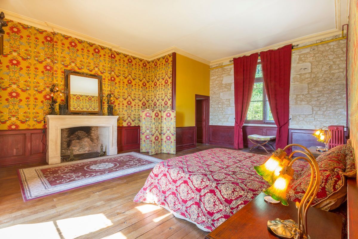 Une pièce avec une cheminée, un lit recouvert d’une couvre-lit à motifs rouges et blancs, un plancher en bois et un mur en pierre.