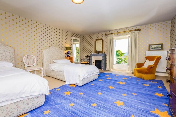 A bedroom with two beds, a fireplace, a yellow armchair, and a view of greenery outside.