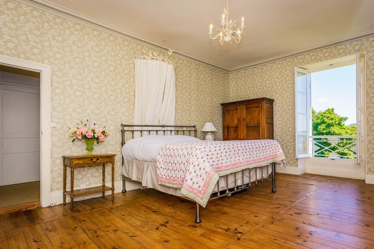 A bedroom with a wooden floor, a bed with a floral bedspread, a nightstand with a vase of flowers, a wooden wardrobe, and a window with a view of greenery.