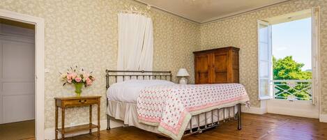 A bedroom with a wooden floor, a bed with a floral bedspread, a nightstand with a vase of flowers, a wooden wardrobe, and a window with a view of greenery.