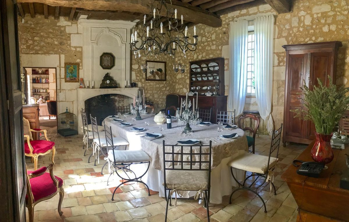 Un comedor con una mesa redonda dispuesta para cuatro personas, rodeada de sillas. Hay una chimenea con repisa, una araña y una estantería con diversos objetos.