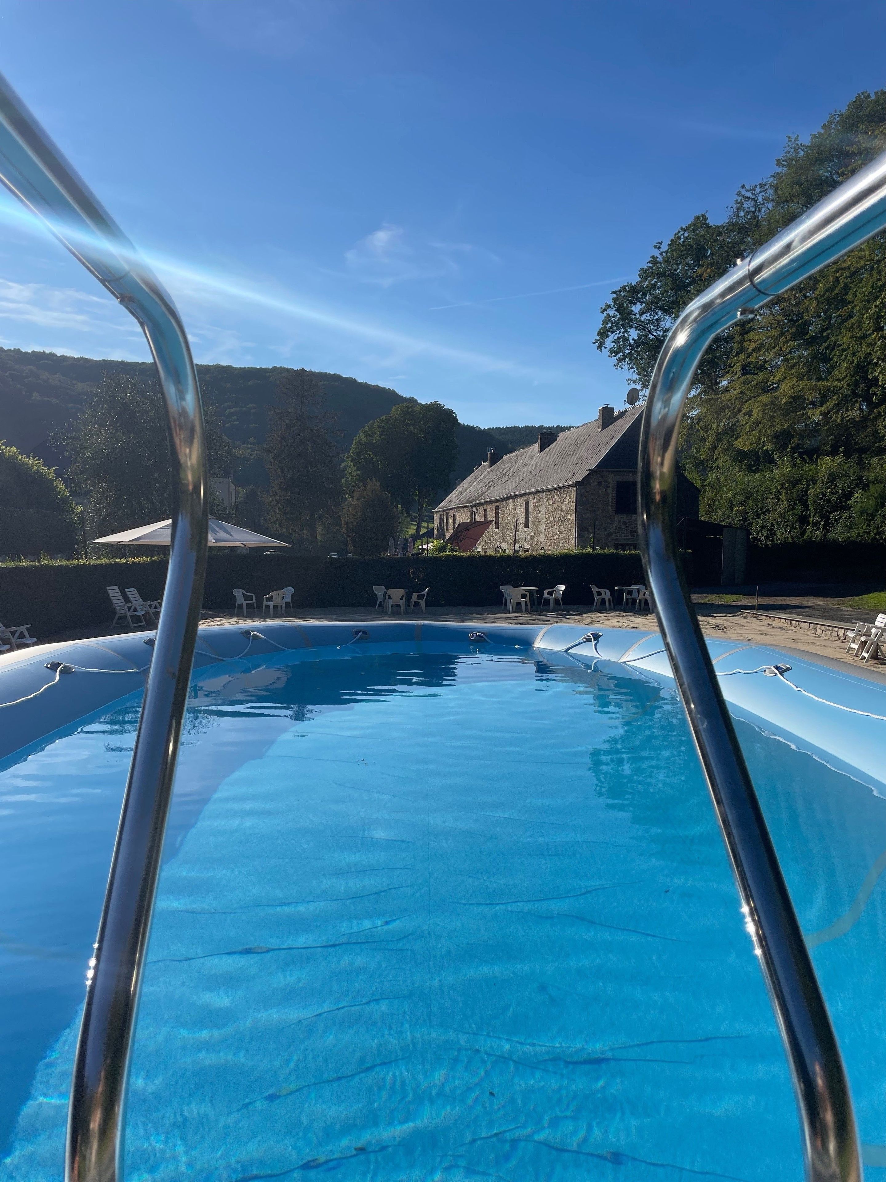 Une piscine avec un escalier en métal au premier plan, entourée de chaises longues et d’un bâtiment en pierre en arrière-plan.