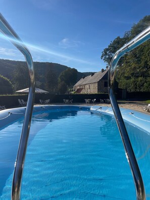 A swimming pool with a metal ladder in the foreground, surrounded by lounge chairs and a stone building in the background.