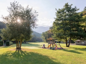 Ein Park mit Rasen, einer Spielplatzanlage, Bäumen und einer entfernten Hügellandschaft.