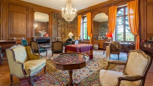 Une vaste pièce aux boiseries richement décorées, agrémentée d’un lustre et dotée d’une grande fenêtre ornée de rideaux orange.
