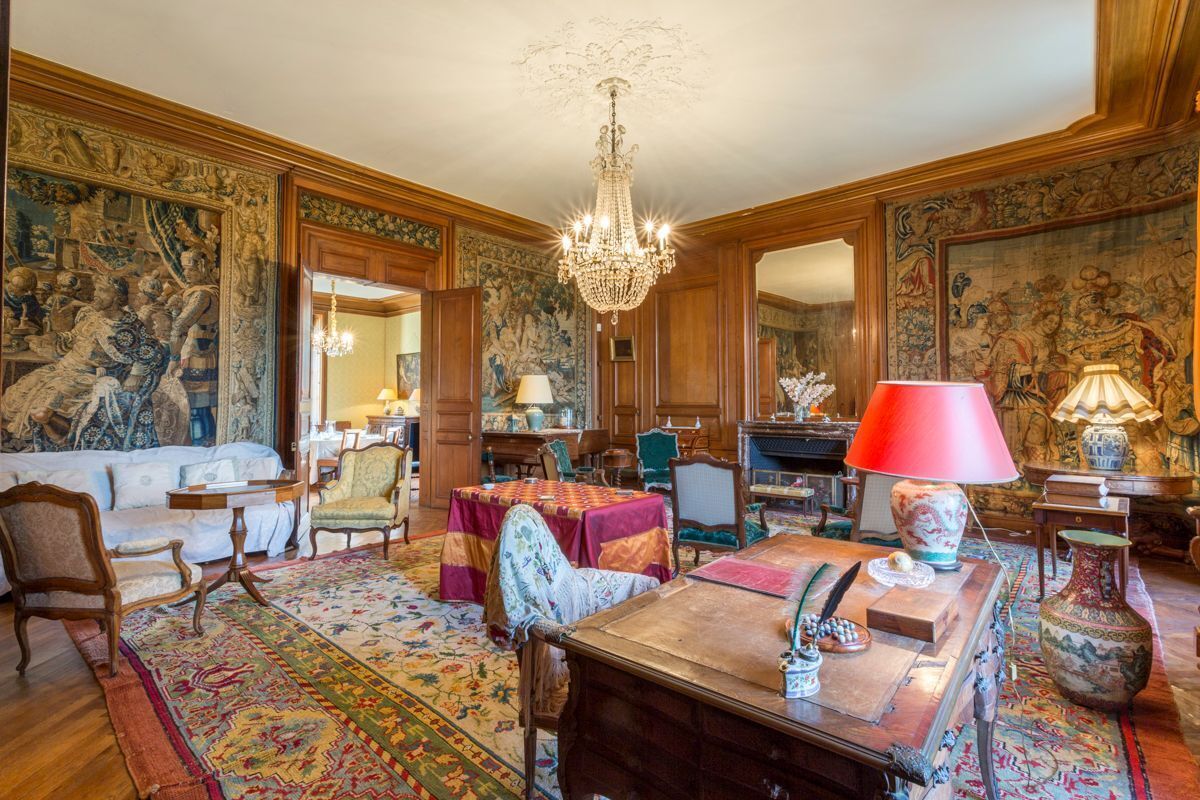 Een klassieke kamer met houten vloer, een kroonluchter, een bank, een tafel met rode lamp en een groot wandtapijt.