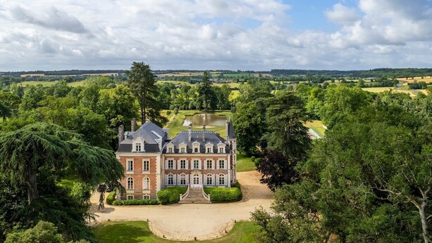 Un grand bâtiment historique à la façade symétrique, entouré d’une végétation luxuriante et d’une pelouse bien entretenue.