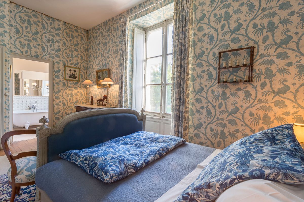 A bedroom with floral wallpaper, a bed with blue bedding, a wooden chair, a dresser, a mirror, and a window with curtains.
