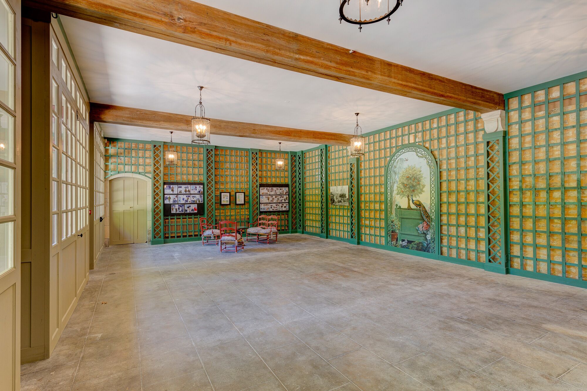 A spacious room with a high ceiling, wooden beams, and a green patterned wall. There are two chandeliers, a wooden bench, and a door with glass panels.