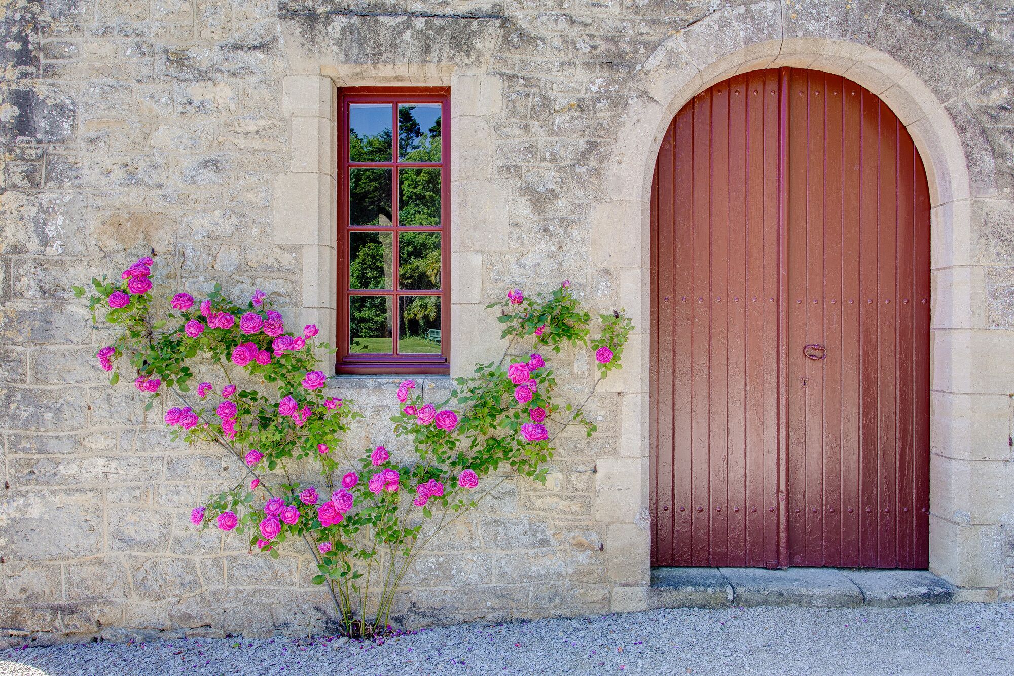 A stone building with a large arched wooden door and a window with a view of greenery.
