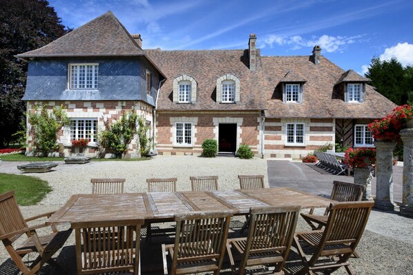 Une maison à deux étages avec une façade en pierre, une allée gravillonnée et, au premier plan, une table et des chaises en bois.