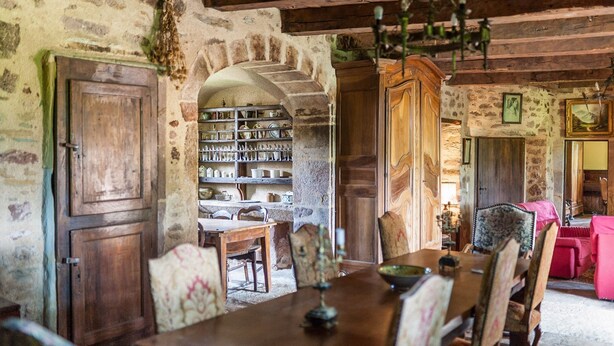 A rustic dining room with a wooden table and chairs, a stone archway leading to a kitchen, and a cozy living area with a pink sofa.