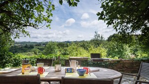 An outdoor dining area with a view of a lush valley, a stone wall, and a wooden table set with various dishes and a kettle.