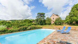 A swimming pool with stone flooring, surrounded by greenery and a stone building in the background.