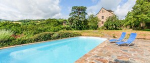 A swimming pool with lounge chairs, surrounded by greenery and a stone building in the background.