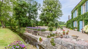 Ein Garten mit einem schmalen Wasserlauf, Steinmauern und verschiedenen Kübelpflanzen.