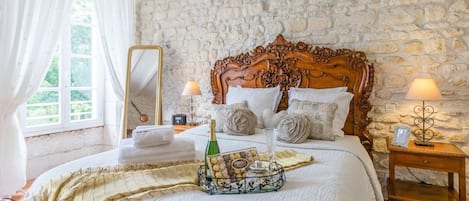 A bedroom with a stone wall, a wooden bed frame, a bedside table with a lamp, and a window with curtains.