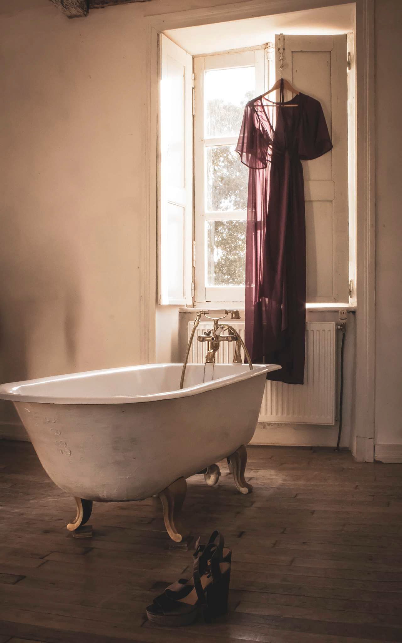 A vintage clawfoot bathtub in a room with a wooden floor, a window with a view of trees, and a hanging garment.