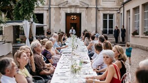 Una larga mesa de comedor adornada con manteles blancos y centros de mesa florales, rodeada de invitados sentados en un patio exterior con arquitectura clásica.