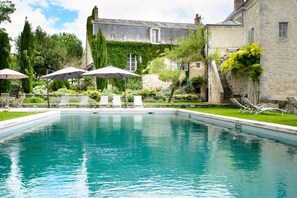 Una piscina rectangular con sillas de descanso y sombrillas, rodeada de exuberante vegetación y edificios de piedra.