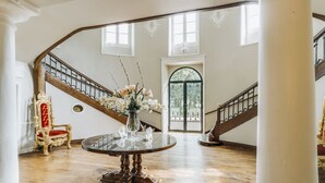 Un gran salón con una escalera de madera, una silla grande y ornamentada, una mesa redonda con un jarrón de flores y un amplio arco de entrada.