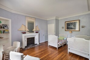 Ein Zimmer mit Kamin, zwei weißen Betten, einer Bücherablage und einem gerahmten Bild an der Wand.