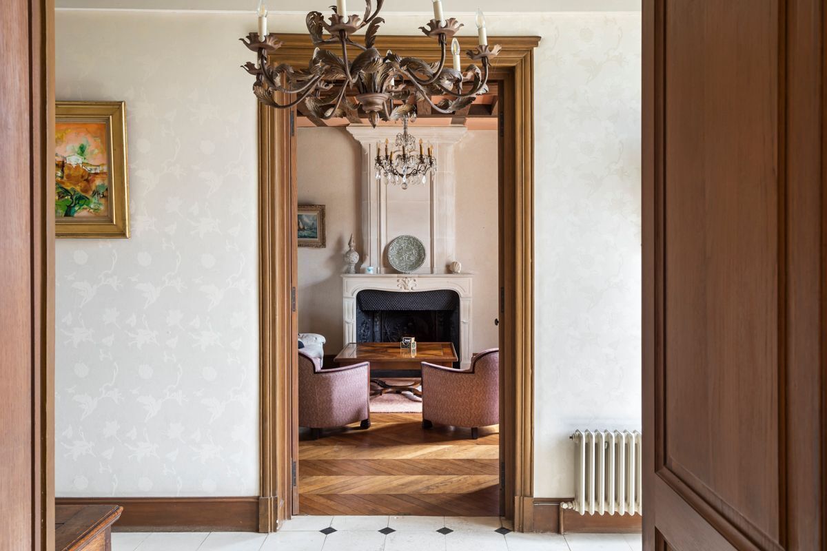 A room with a fireplace, two armchairs, a wooden floor, and a chandelier.