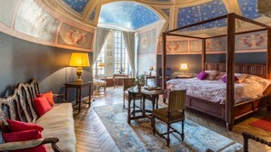 A four-poster bed with a floral bedspread, a wooden bench, a table with chairs, and a sofa with red cushions in a room with a high, vaulted ceiling and floral wallpaper.