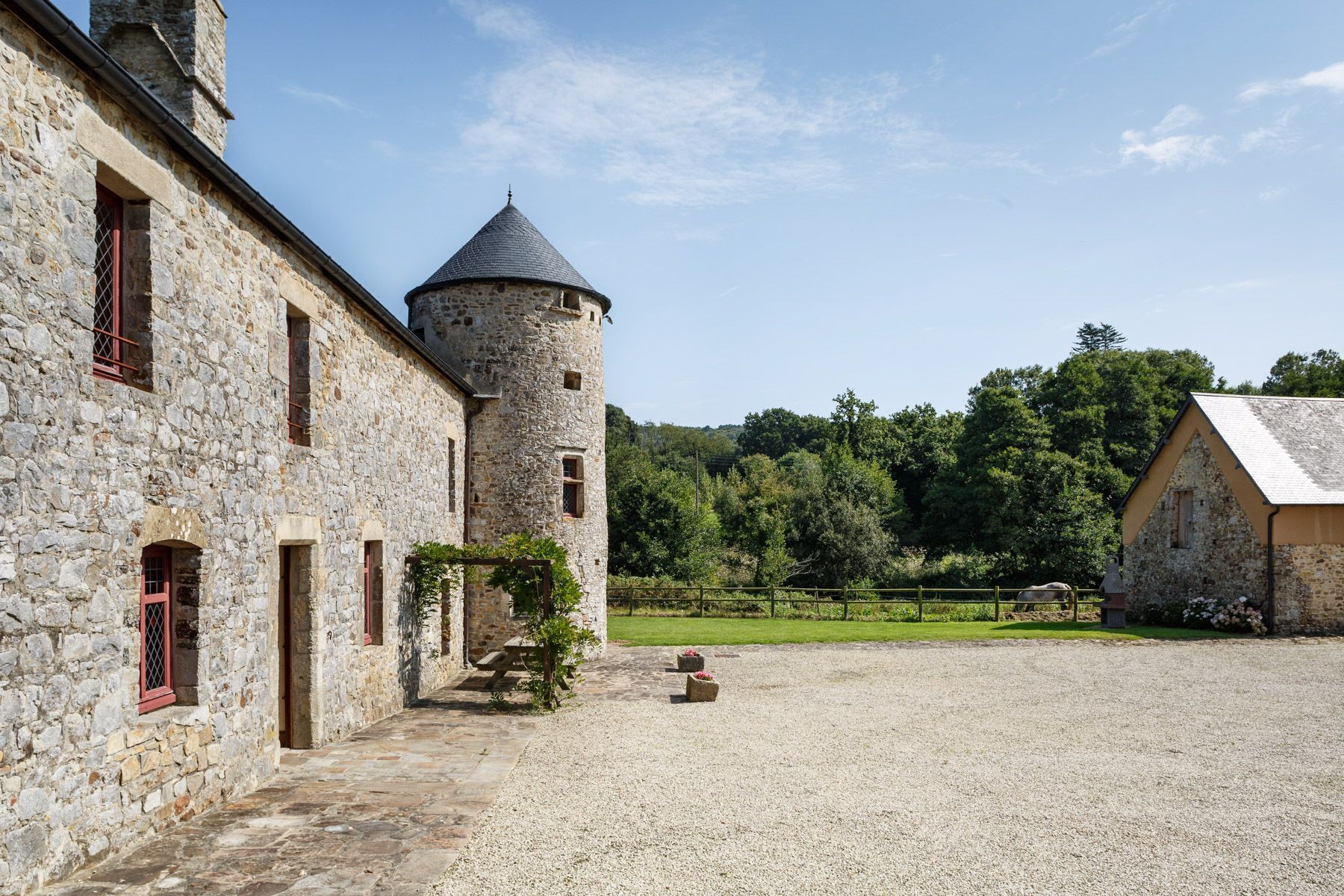 Un bâtiment en pierre doté d’une tour, une cour recouverte de gravier et, au loin, une colline boisée.