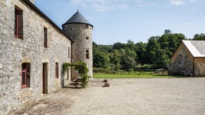 Un edificio de piedra con una torre, un patio de grava y una colina boscosa al fondo.