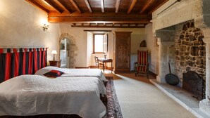 Una habitación tradicional con chimenea de piedra, dos camas con sábanas blancas, techo de madera y pared de piedra.
