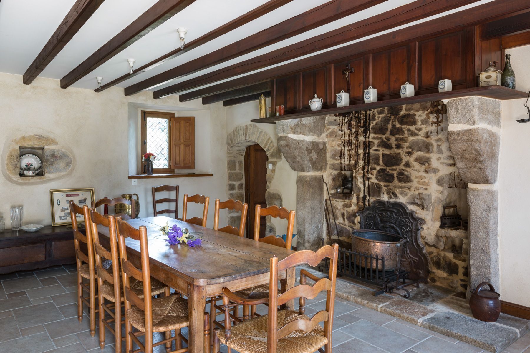 Un espace repas agrémenté d’une cheminée en pierre, de poutres apparentes et d’un sol carrelé.