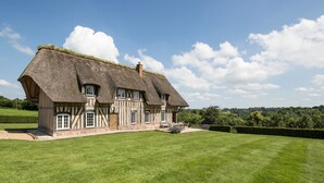 Une maison traditionnelle à colombages, avec un toit de chaume, une pelouse bien entretenue et un ciel bleu dégagé.