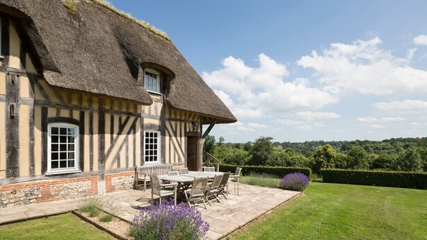 Une maison traditionnelle à colombages, avec un toit de chaume, une terrasse avec une table et des chaises, et un jardin bien entretenu.