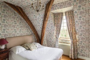 A bedroom with a wooden ceiling, patterned wallpaper, a bed with white linens, a nightstand with a lamp, and a window with curtains. - Domaine Du Pont: Entire Estate (Saint-Maclou)