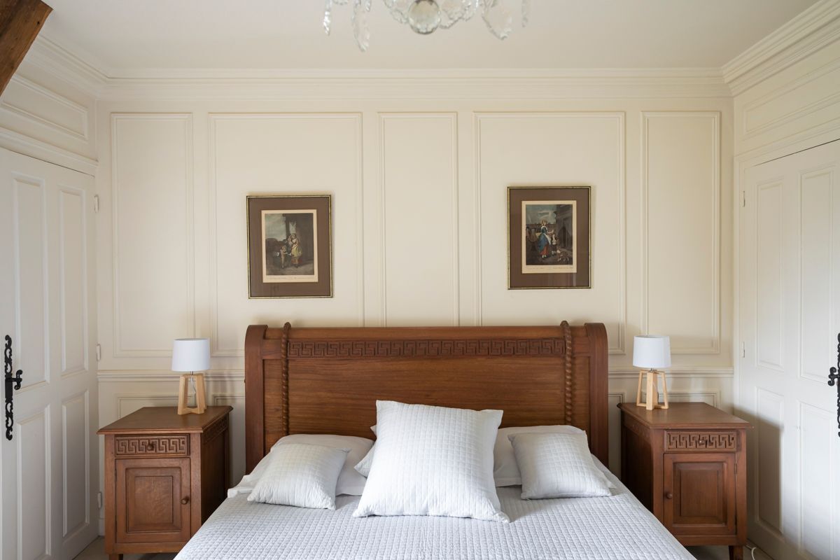 Une chambre à coucher avec une tête de lit en bois, deux tables de chevet avec des lampes, un lit avec une literie blanche et deux tableaux encadrés accrochés au mur.