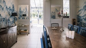A dining area with a long table, blue chairs, and a chandelier. There are paintings on the walls and a window with a view of greenery.