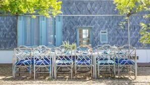 A covered outdoor seating area with a table and chairs, a window with shutters, and a tree in the background.