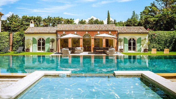 A pool with a clear water surface, a patio area with outdoor furniture, and a building with a tiled roof and arched windows.