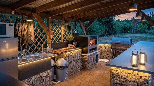 Une cuisine extérieure avec un mur en pierre, un barbecue, un évier et un téléviseur.
