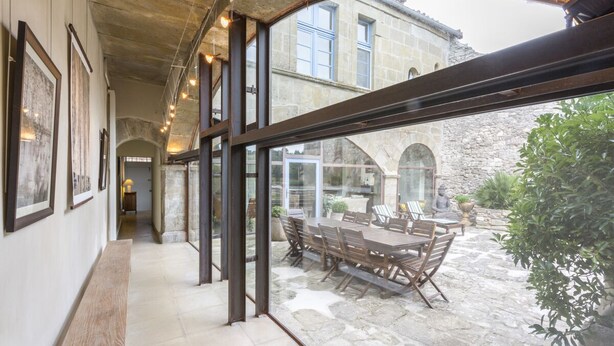 A corridor with framed artwork on the walls, a wooden bench, and a glass door leading to an outdoor patio with a table and chairs.