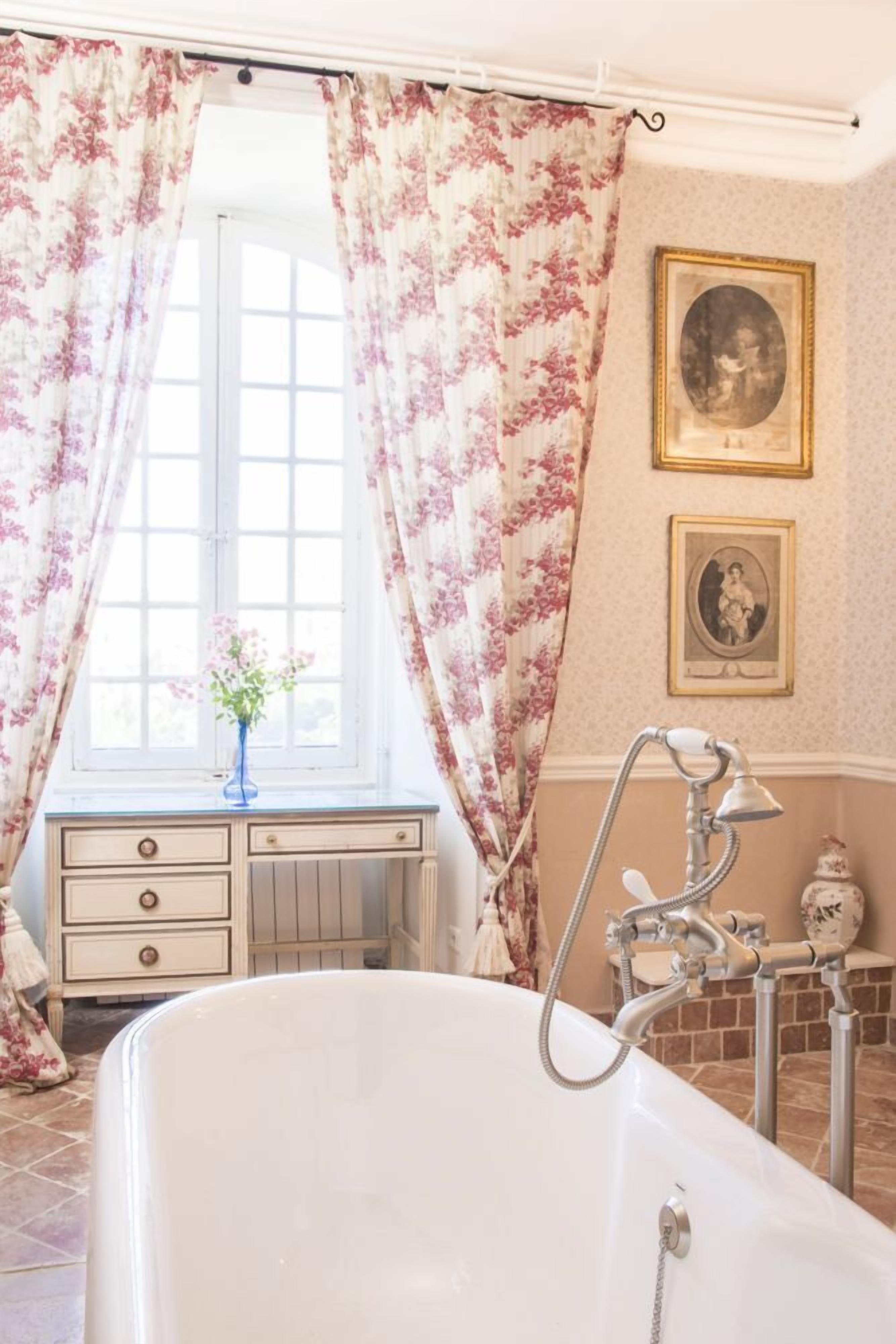 A bathroom with a bathtub, floral curtains, a window, a dresser, and framed pictures on the wall.